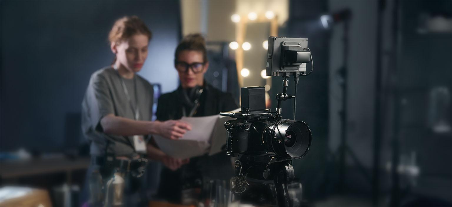  Cámara de video en primer plano con dos personas de fondo revisando documentos en un set de filmación.